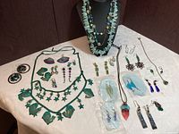 Full overview of the laid out costume jewelry including necklaces and earrings featuring green, blue, and purple colors with various materials such as patina metal, polished stones, and glass beads.