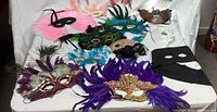 Various feathered and sequin costume masks displayed on white cloth, showing colors purple, black, blue, and gold trimming.