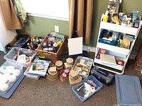 Photo showing an overview of boxed and loose craft supplies including bottles of paint, glue, boxes, and a white metal cart filled with various tools