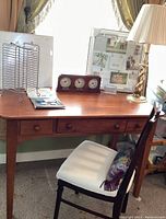 Wooden desk with single drawer, chair with white cushion and pillow, brass desk lamp, analog clock with three dials, metal file organizer, and whiteboard with dry erase markers arranged on desk in front of window.
