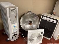Full view of all four heating and cooling appliances: DeLonghi oil-filled ceramic heater, HeatDish heat lamp, Safe & Solid electric heater, and small window fan.