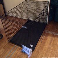 Large black metal dog crate with plastic tray base and attached door, with plastic bag of instructions and handles on front.
