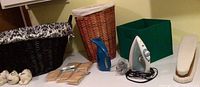 Laundry items on table including dark wicker hamper, medium brown wicker hamper, green fabric bin, blue fabric steamer, electric iron, and wooden clothespins.