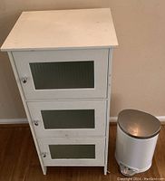 Front view of white wooden cabinet with frosted ribbed glass doors and white metal pedal trash can beside it.