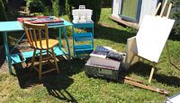 Overall outdoor view of turquoise craft table with wood chair, white organizer baskets on cart, easel with blank canvas and art supplies on the table.