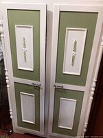 Front view of white and olive hand-painted cabinet doors with carved floral vertical panels and handles.