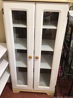 Front view of white painted pressed wood cabinet with two glass insert doors and wooden knobs, standing on floor.