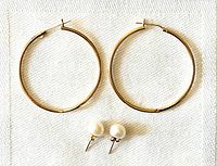 Pair of 14K yellow gold hoop earrings and pair of natural freshwater pearl stud earrings laid flat on white textured fabric.
