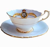 Side view of the white porcelain teacup with blue and gold RCAF insignia and gold-rimmed scalloped edge, seated on matching saucer.