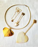 Three costume jewelry necklaces displayed on a white fabric with a gold tone leaf pendant, small pearl heart pendant, and large iridescent heart pendant laid out.