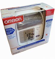 Front and side view of the Omron BP765CAN blood pressure monitor in the sealed retail box showing the device and packaging details.