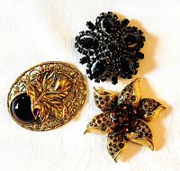 Photo showing three vintage brooches laying flat: oval gold-tone leaf and black stone brooch, black rhinestone floral brooch, gold-tone flower brooch with red stones.