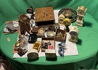 Wide shot of the full lot showing multiple antique and vintage alarm clocks and assorted clock parts on a white surface with green background.