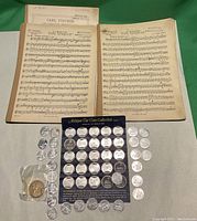 Photo showing Franklin Mint antique car coin collection card with 32 visible silver-tone coins, multiple loose silver-tone coins, one loose gold-toned coin in plastic sleeve, and two sheets of vintage music notes by Carl Fischer.