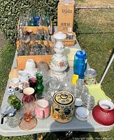 Full view of the large lot displayed on a table outdoors showing antique glass bottles, a large white floral glass drink dispenser, various colored glassware, ceramics, and a decorative tin container.