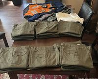 Neatly folded stacks of olive drab army work pants in various sizes along with several folded upper body garments including jackets and a white and an orange reflective vest