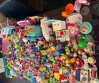 Large assortment of various kids toys spread out on a table including dolls, plush animals, building blocks, and kitchen play items.