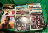 Photo showing 19 LaserDisc movie cases and sleeves arranged on a green surface. Titles like GoldenEye, Die Hard with a Vengeance, Rush, and The First Knight are visible.
