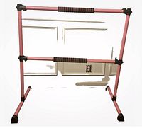 Front view showing pink adjustable portable ballet barre with dual horizontal bars, black foam grips, and black rubber feet on wooden floor against white wall.