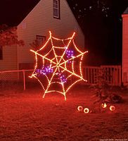 Spider web Halloween decoration lit up outdoors at night, showing orange LED lights outlining the web and purple LED lights on spiders attached to the web.