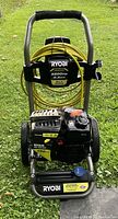Full view of Ryobi pressure washer showing yellow high pressure hose wrapped around handle, black engine with Kohler label, and metal frame with wheels.