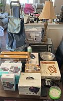 Group shot of mixed kitchen and home items including shopping case, toaster oven, phone sanitizer box, security camera, wine rack, table lamp, kitchen bowls and packaged accessories.