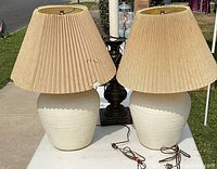 Two vintage pottery table lamps with cream ceramic bases and beige pleated lampshades placed side by side outdoors on a white table.