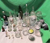 Full view of assortment of vintage bottles and jars arranged on white surface with green backdrop. Includes multiple clear glass jars with wire bale lids, dark glass bottles, seltzer bottle with metal spout, small vials, and several vintage Coca-Cola bottles with embossed logos. Stack of coiled metal lids on right.