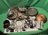 Wide view of the assortment of vintage kitchenware items on green backdrop including meat grinder, metal and brass plates, utensils, and copper lid.