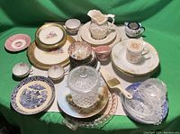 Wide view of lot items showing assorted porcelain plates, pitcher, cups, and glassware on green cloth background.