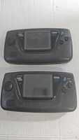 Top view showing two black Sega Game Gear handheld consoles side by side with visible screens and button layouts. Both show wear marks.