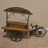Side angle view of the wooden bicycle wagon planter showing the front wheel, wagon bed with an open lid, and the 'FLOWERS and PLANTS' sign above.