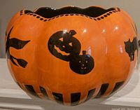 Close up photo of orange ceramic Halloween punch bowl showing black cat, jack-o-lantern faces and bat motifs with scalloped rim detail and striped base.