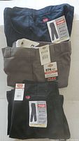 Three pairs of folded work pants stacked, with tags visible showing Dickies and Wrangler brands, two pairs black and one tan.