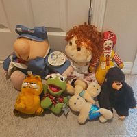 Group photo showing entire lot of 1980's plush toys including Garfield, Kermit, postal worker, red-haired doll, Ronald McDonald, two mermaids, small turtle and gorilla.
