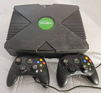 Original Xbox console on a white surface with two wired controllers placed in front.