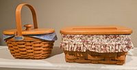 Two vintage Longaberger baskets shown side by side, one with handle and blue patterned liner, other rectangular with floral ruffled liner