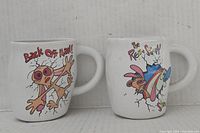 Shows front view of both mugs side by side with their cartoon graphics visible.