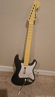 Black Fender Stratocaster style guitar controller with a cream-colored fretboard and white pickguard against a beige wall on carpet.