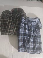 Two folded plaid button-up shirts laid side by side on flat surface, one Tommy Hilfiger and one Ralph Lauren.