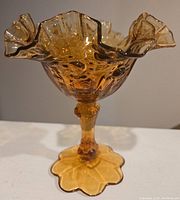 Side view of a vintage amber glass compote showing hobnail pattern on bowl and stem, ruffled edges, and petal-shaped base.