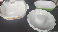 Photo showing all three dishes: clear glass casserole with floral design and lid, green and white rectangular ceramic baking dish, and white ceramic fluted tube pan.