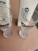 Two Cheers Boston glass bar glasses standing side by side with original boxes behind them.