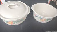 Two Corelle stoneware cooking pots on black surface; one pot with lid, one pot without lid. Both feature vegetable-themed decorative pattern.