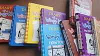 Photo showing front covers of multiple Diary of a Wimpy Kid books arranged in a fan style, displaying colorful and illustrated covers.