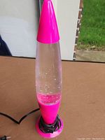 Photo of entire lava lamp standing upright on brown table with black cord wrapped around pink base.
