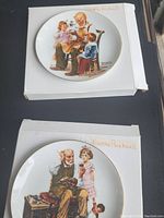 Photo of the two Norman Rockwell decorative plates placed on their respective white cardboard boxes, showing full artwork on each plate.