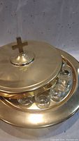 Photo shows the brass lid with a cross handle on top of a brass communion tray with several glass cups beneath the lid.