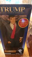 Front view of sealed box showing Donald Trump doll in suit, red tie, and facial features. Box text mentions 12 inch talking doll.