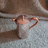 Side view of wicker-handled silver-tone jug, showing spout, handle, and berry-shaped finial on lid.